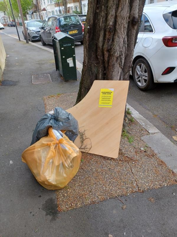 Wooden board and bags containing various items of waste fly tipped underneath a tree at 99 Brock Road, E13. -99 Brock Road, Plaistow, London, E13 8NA